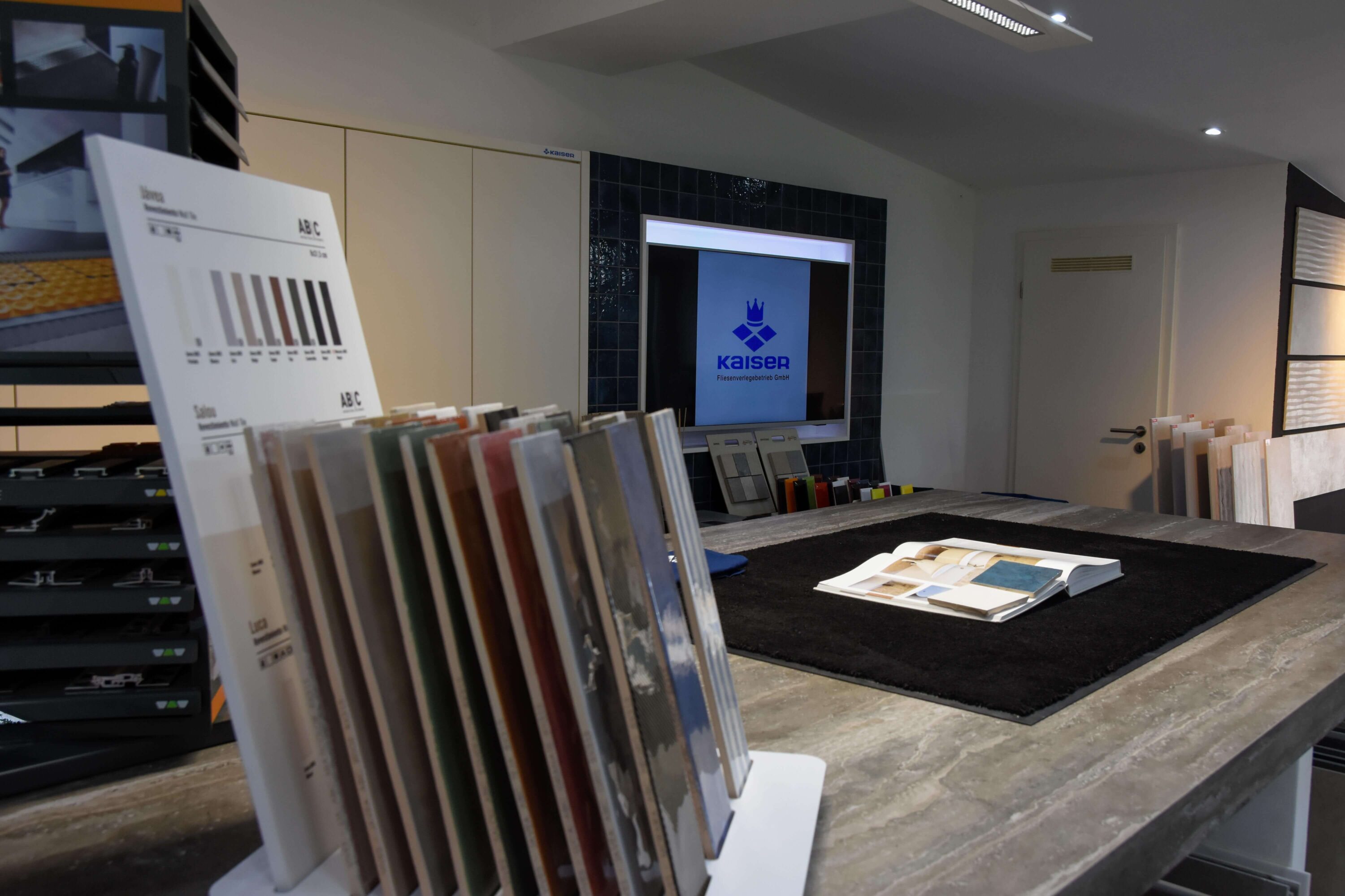 Beratung in der Fliesenausstellung von Kaiser Fliesenverlegebetrieb GmbH. Fliesenausstellung mit Fliesen-Mustern und Fliesen-Katalogen. Im Hintergrund Logo von Kaiser Fliesenverlegebetrieb, Fliesenleger aus Düsseldorf Gerresheim.